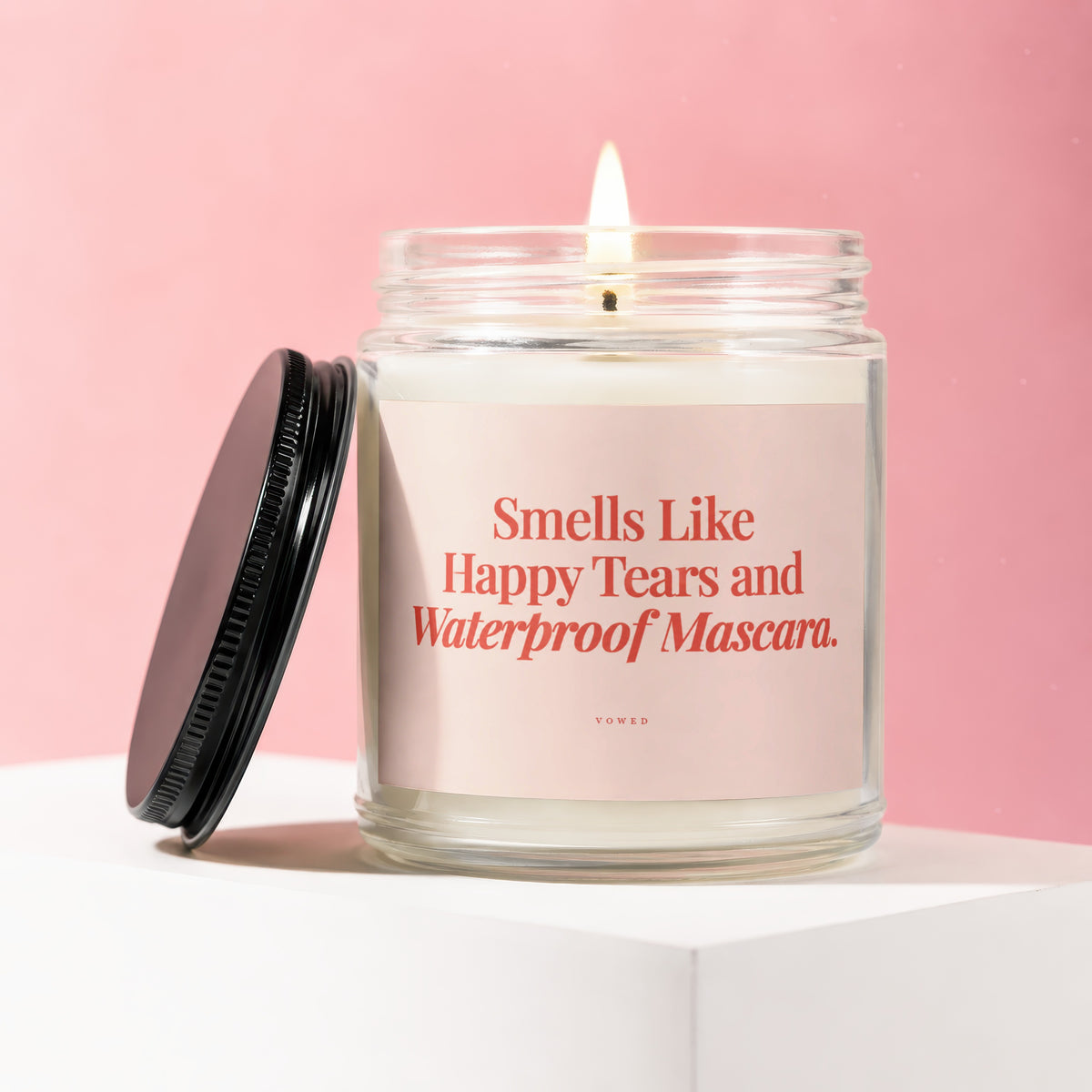 Glass candle with burning wick and black lid, reads "Smells Like Happy Tears and Waterproof Mascara. VOWED".