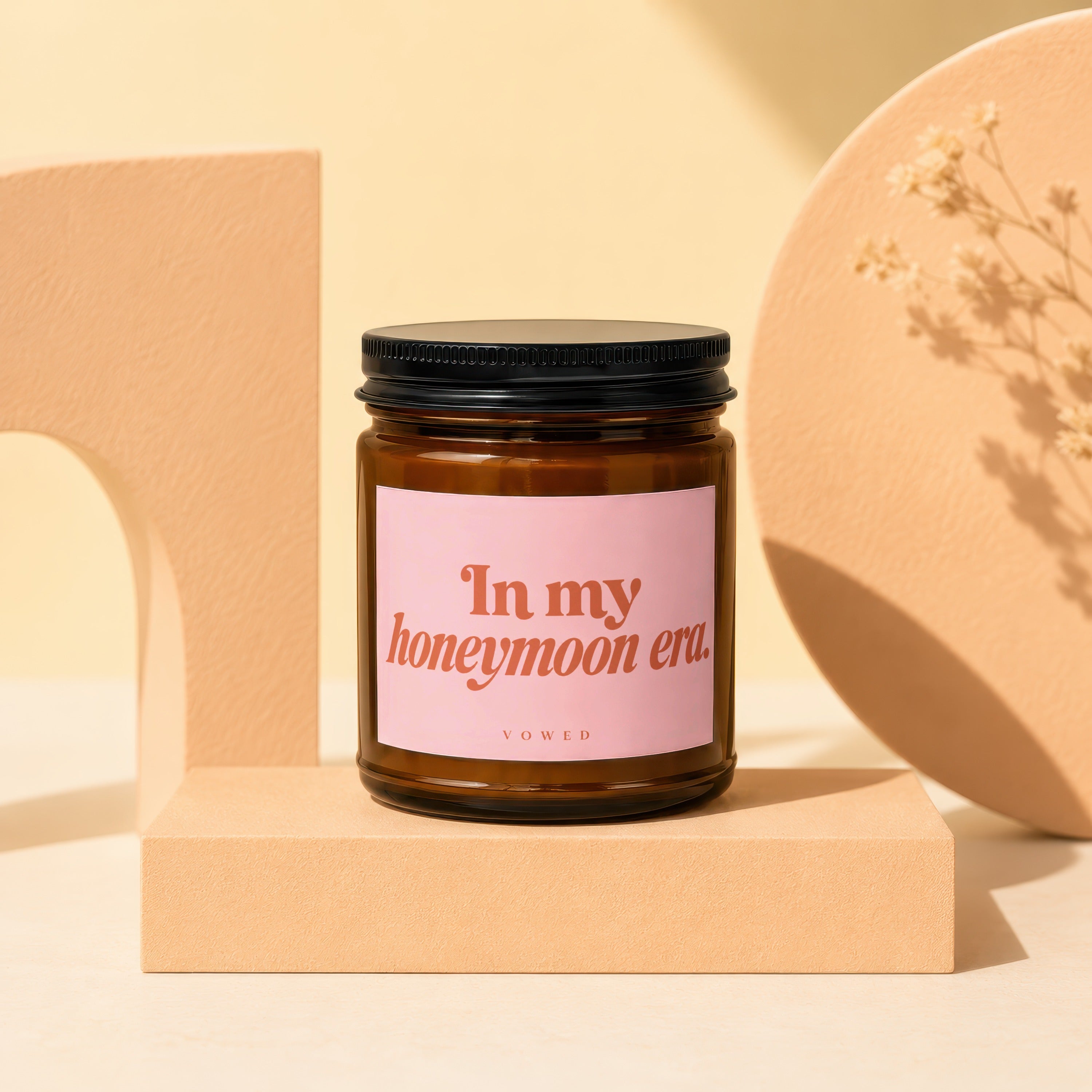 Brown candle in amber glass jar with pink label reading "In my honeymoon era. VO WED."