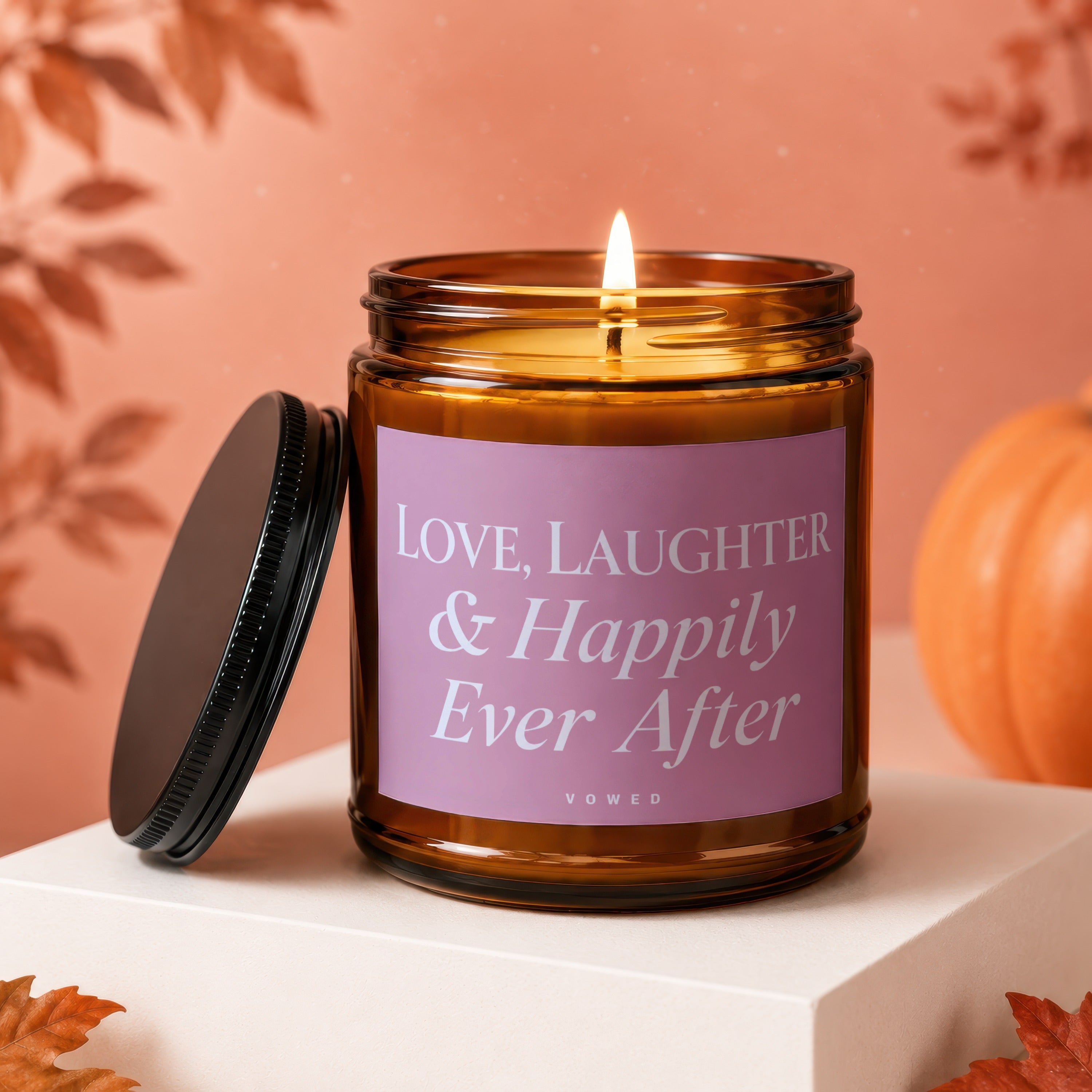 Amber jar candle with a lit wick and a pink label reading "LOVE, LAUGHTER & Happily Ever After".
