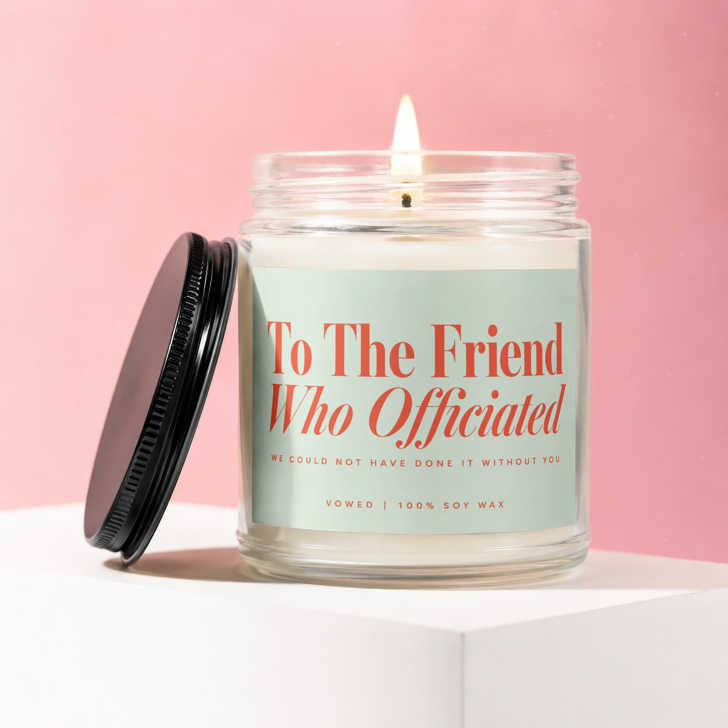 Lit soy candle "To The Friend Who Officiated" on pink background.