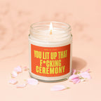 Glass jar candle with lit wick and orange label that reads "You lit up that f*cking ceremony."