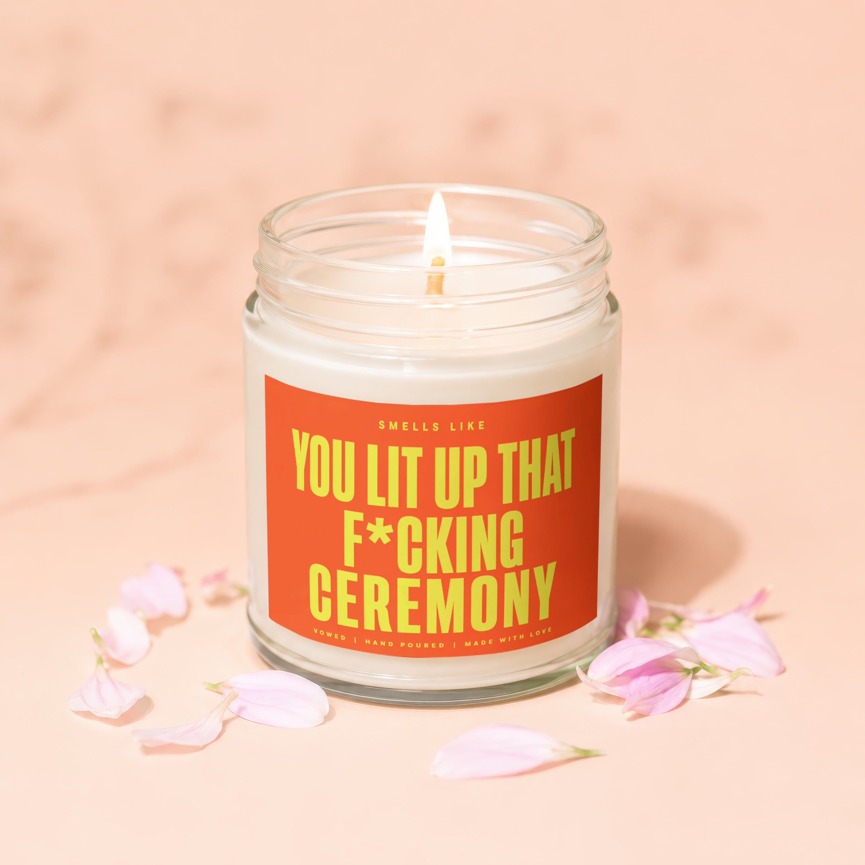 Glass jar candle with lit wick and orange label that reads "You lit up that f*cking ceremony."