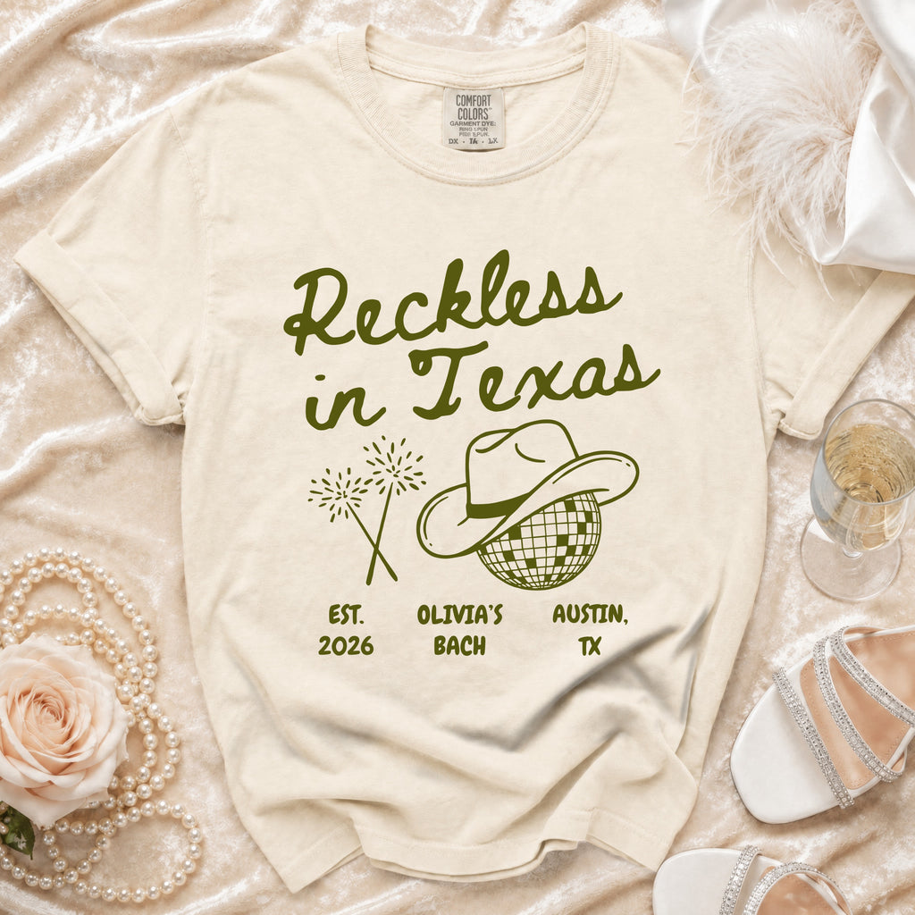 Cream t-shirt with "Reckless in Texas" text, cowboy hat, and fireworks.