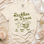 Cream t-shirt with "Reckless in Texas" text, cowboy hat, and fireworks.