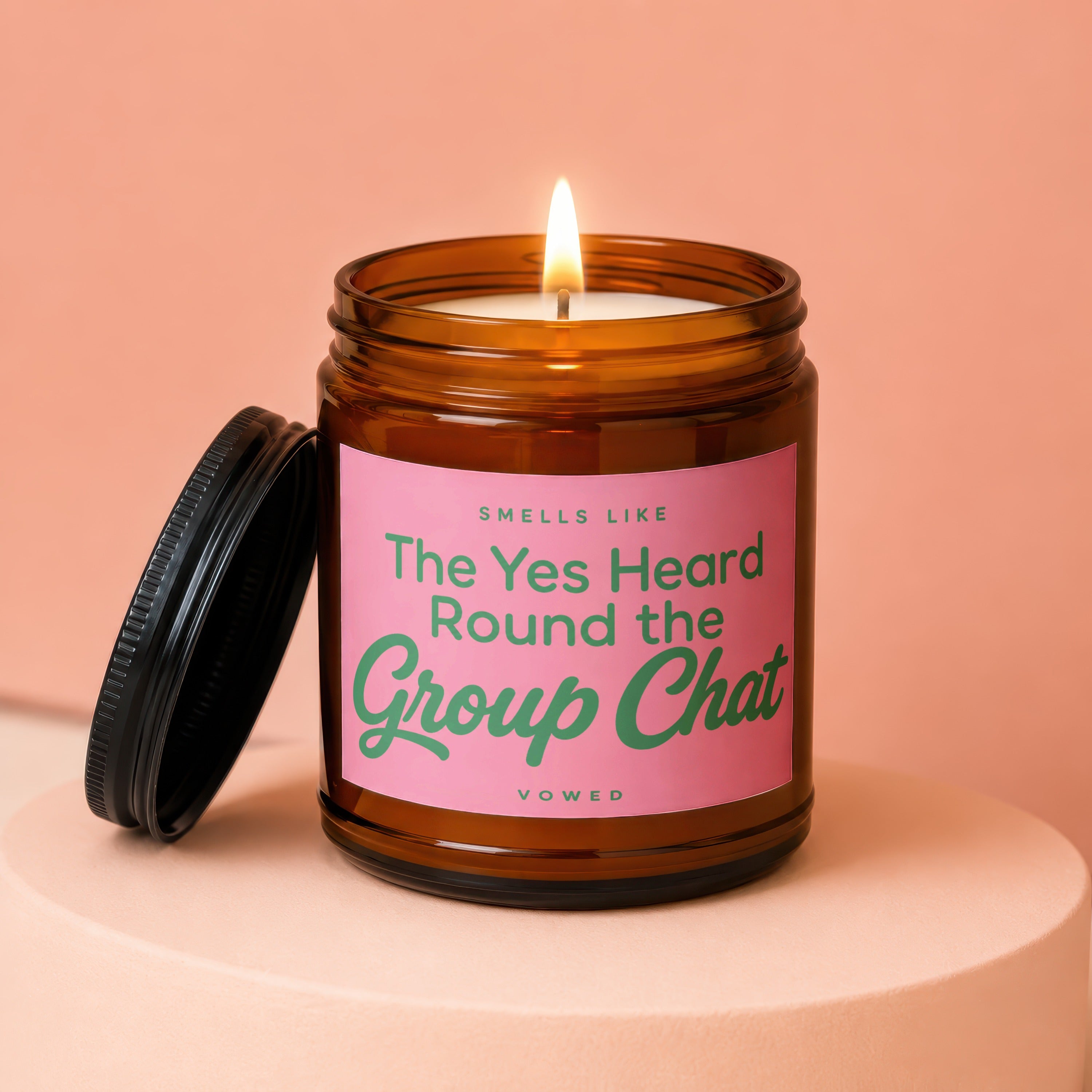 Amber jar candle with lit wick, pink label reads "Smells Like The Yes Heard Round the Group Chat"