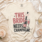 T-shirt with "This Bride Needs Champagne" text, champagne bottle, and glass.