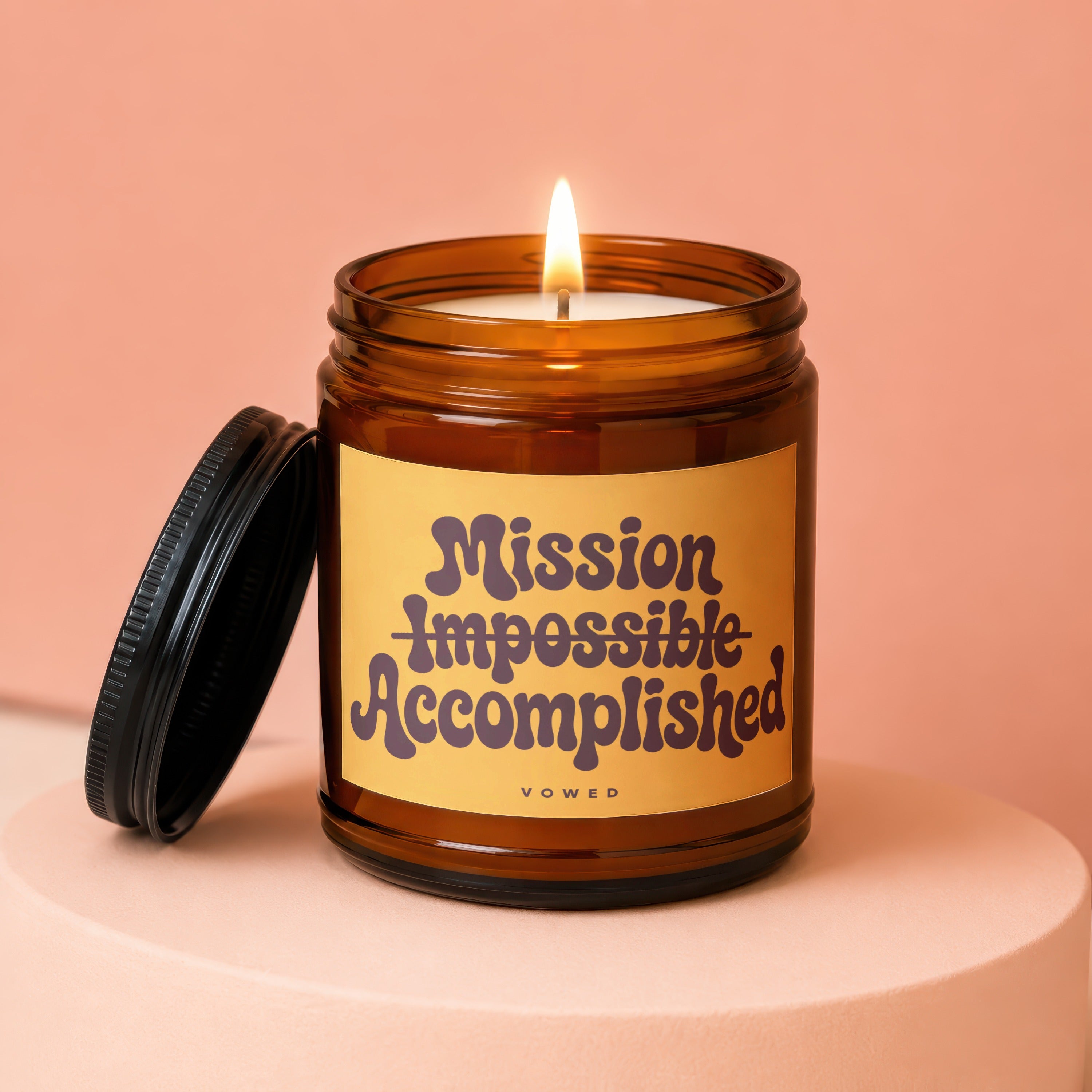 Amber jar candle with lit wick and black lid, reads "Mission Impossible Accomplished".