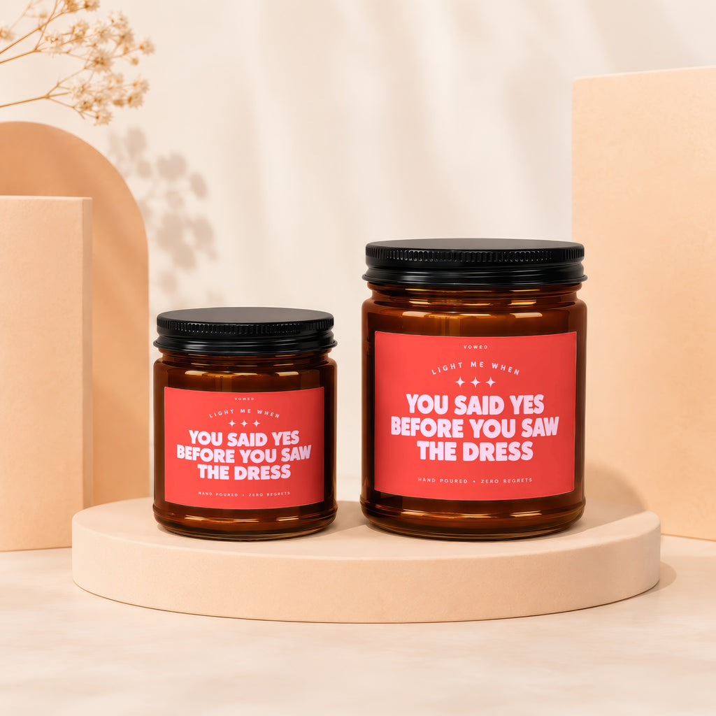 Two amber glass candles with black lids and red labels saying "You said yes before you saw the dress."
