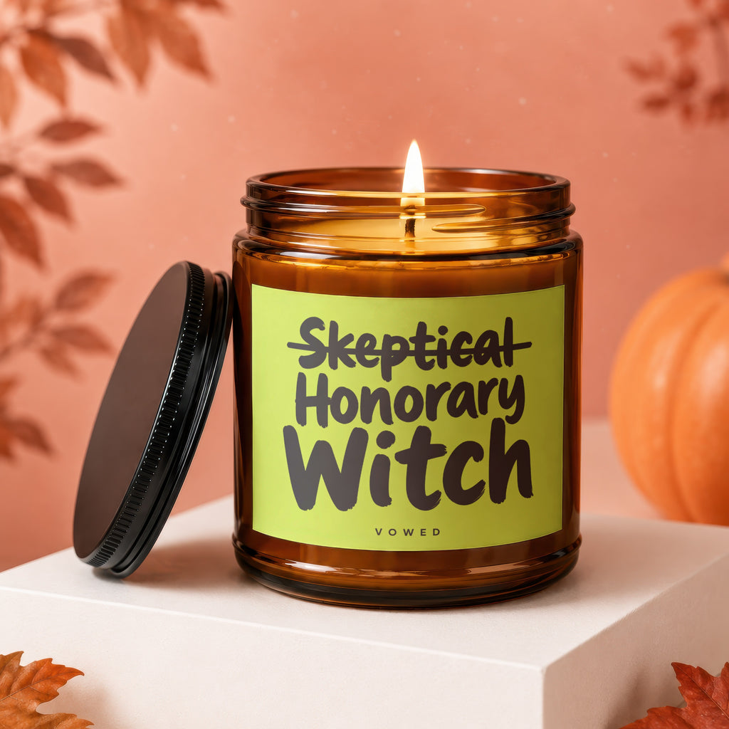 Lit amber glass candle with a green label saying "Skeptical Honorary Witch."
