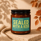 Amber jar candle with "OFFICIAL: SEALED WITH A KISS VOWED" text on teal label.
