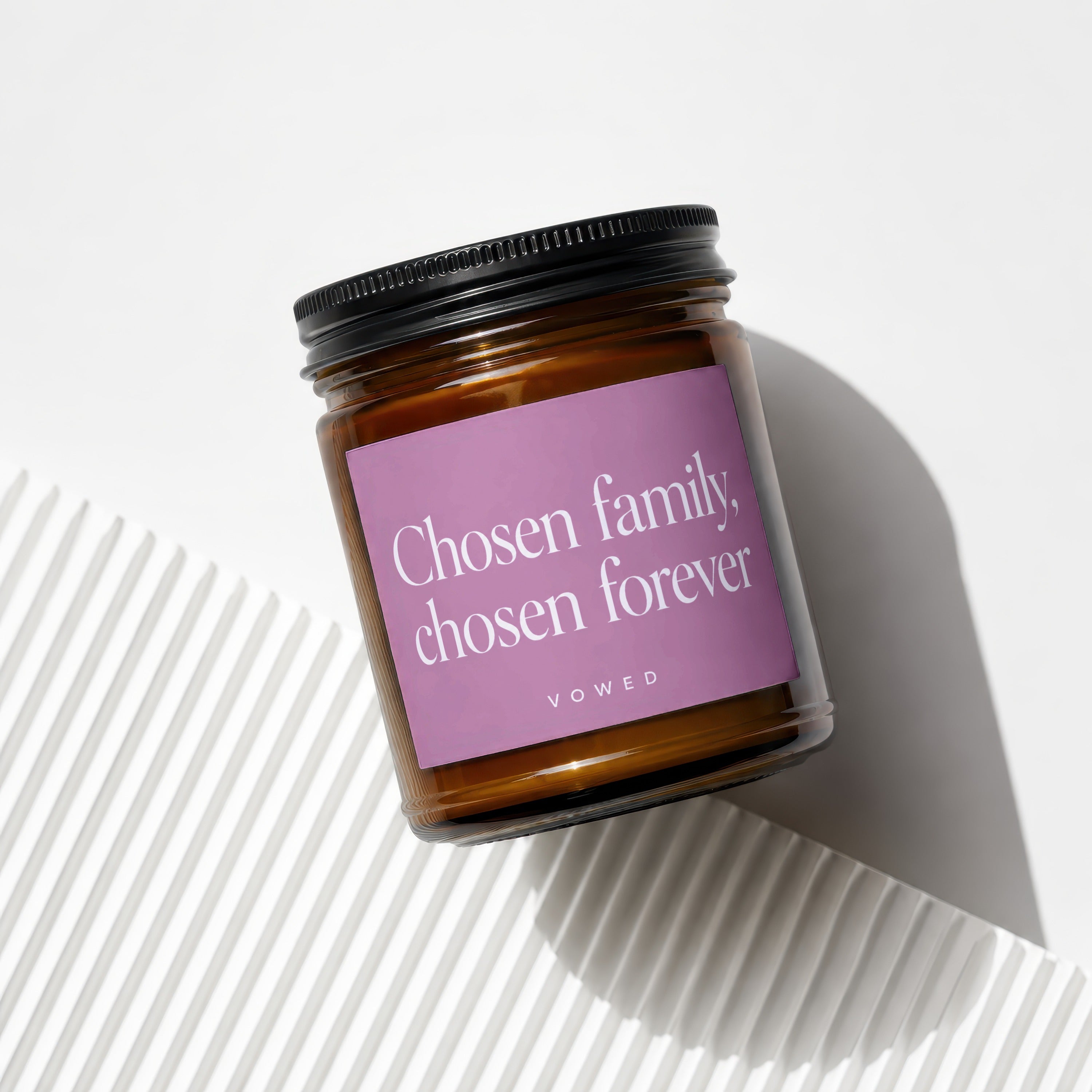 Amber glass candle with black lid and pink label reading "Chosen family, chosen forever."