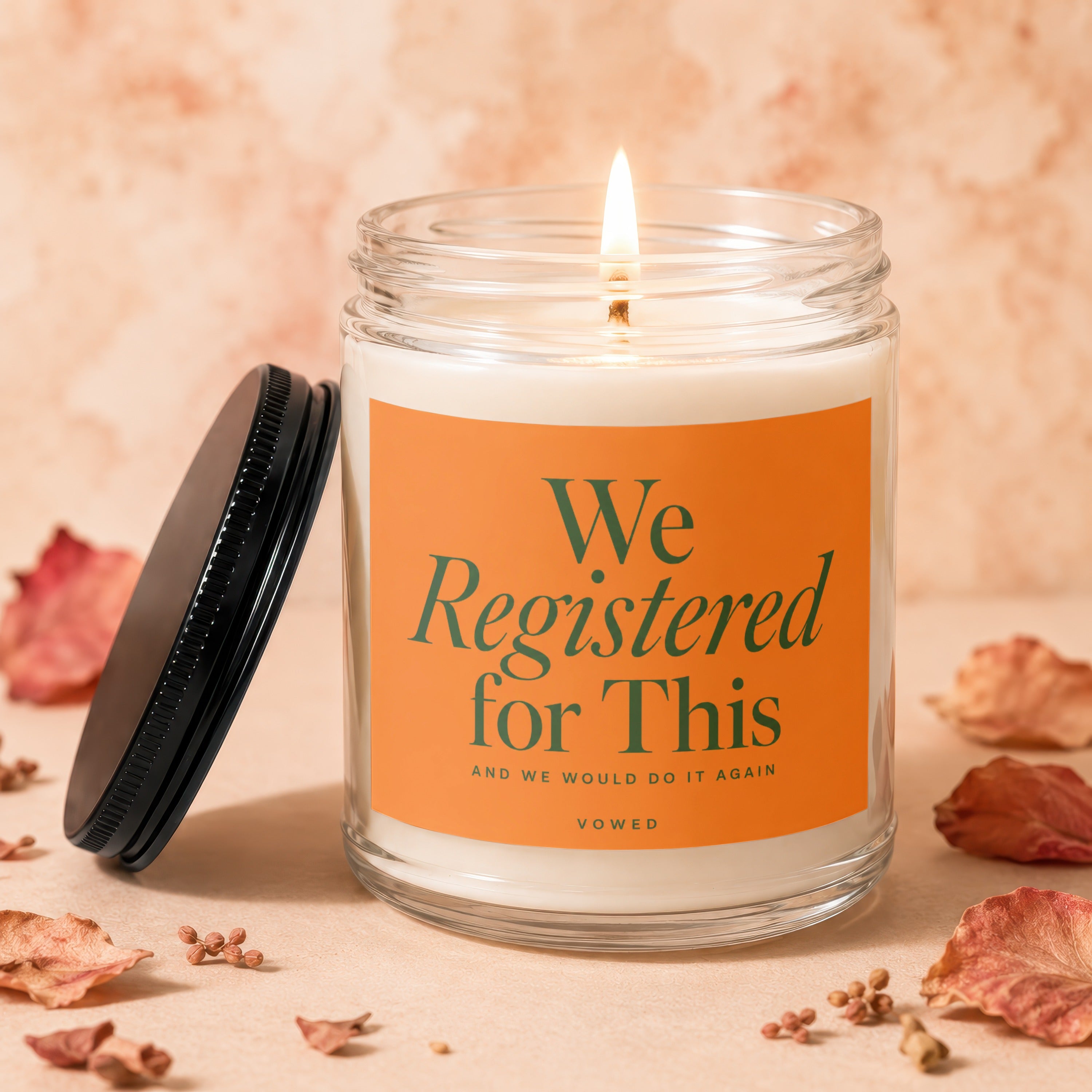 Lit glass candle with orange label reading "We Registered for This" and "Vowed".