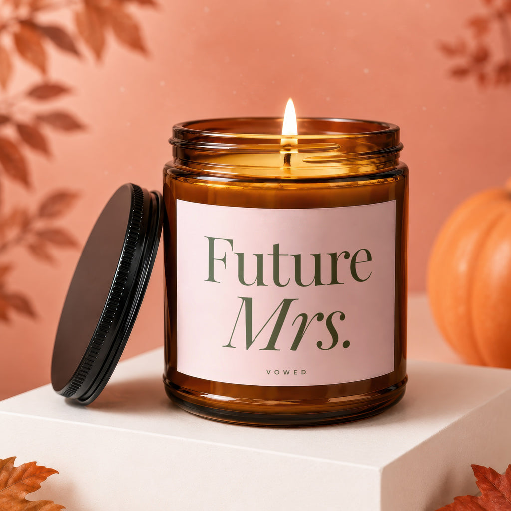 Lit candle with "Future Mrs." text on a pink label, next to its lid, with a pumpkin.