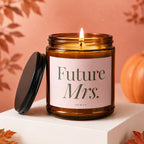 Lit candle with "Future Mrs." text on a pink label, next to its lid, with a pumpkin.