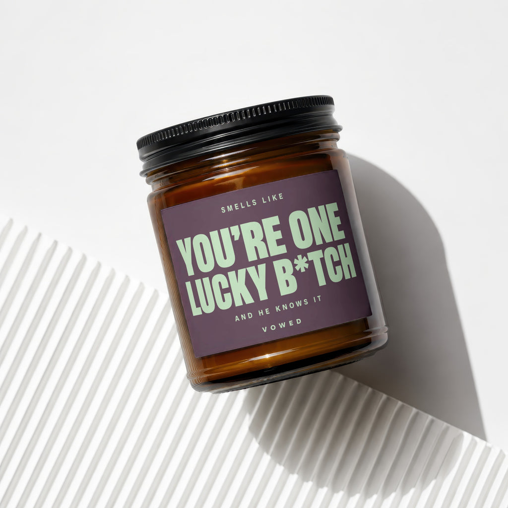 Amber jar candle with dark purple label text "You're One Lucky B*tch And He Knows It".