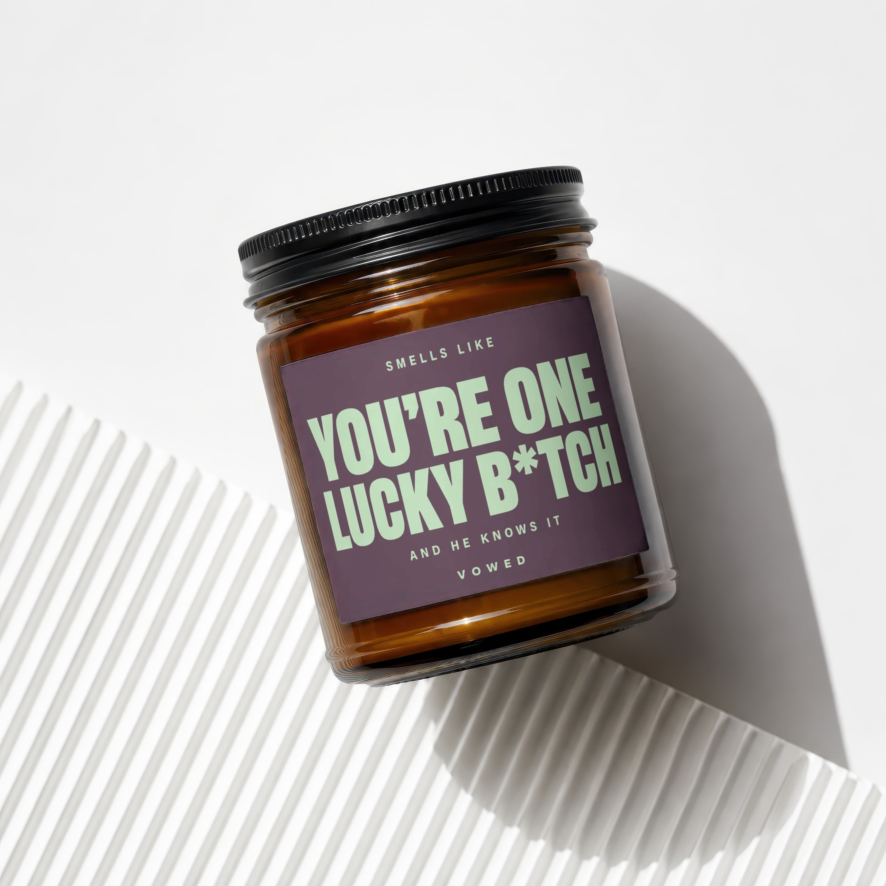 Amber jar candle with dark purple label text "You're One Lucky B*tch And He Knows It".