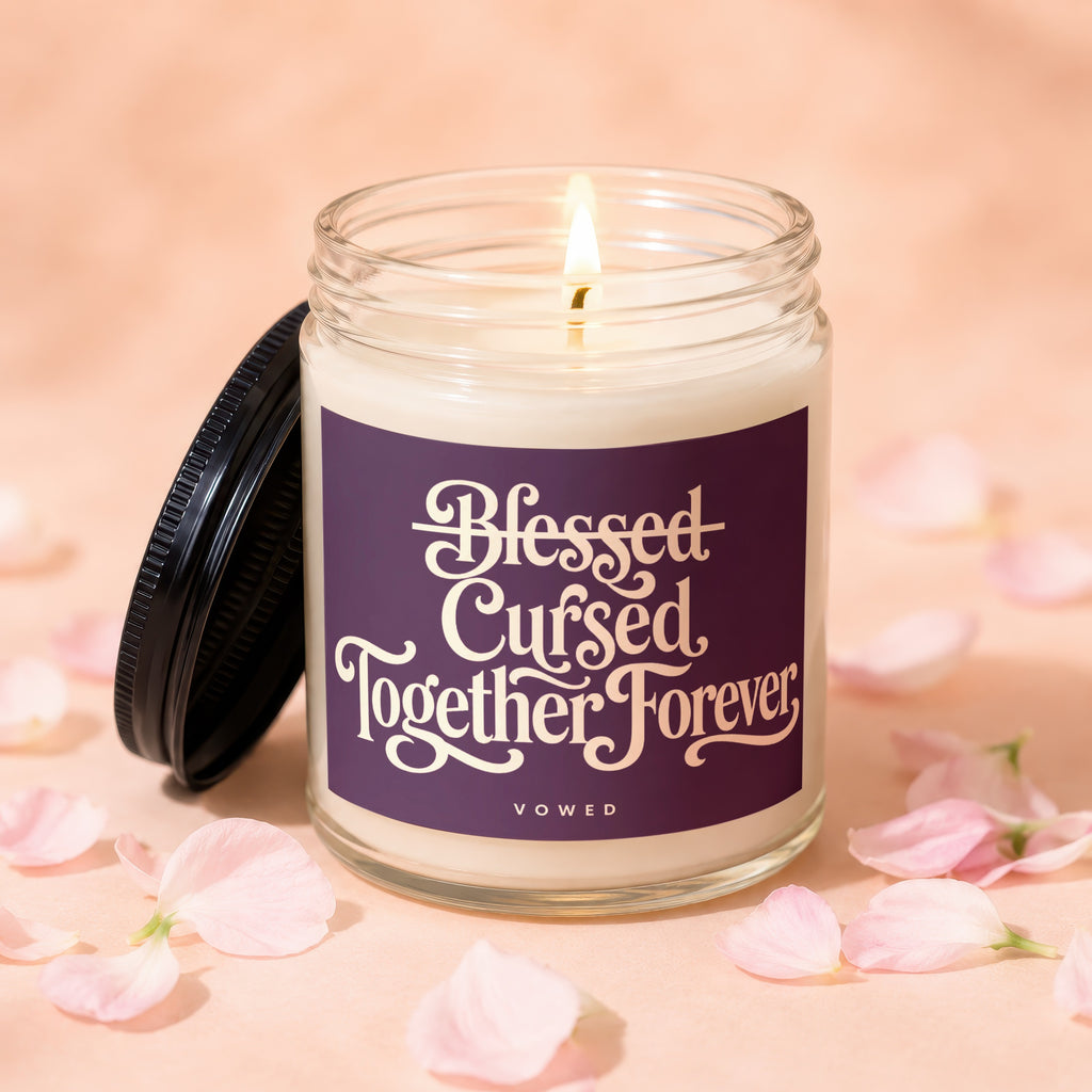 Glass candle with lit wick, black lid, and label reading "Blessed Cursed Together Forever VOWED".