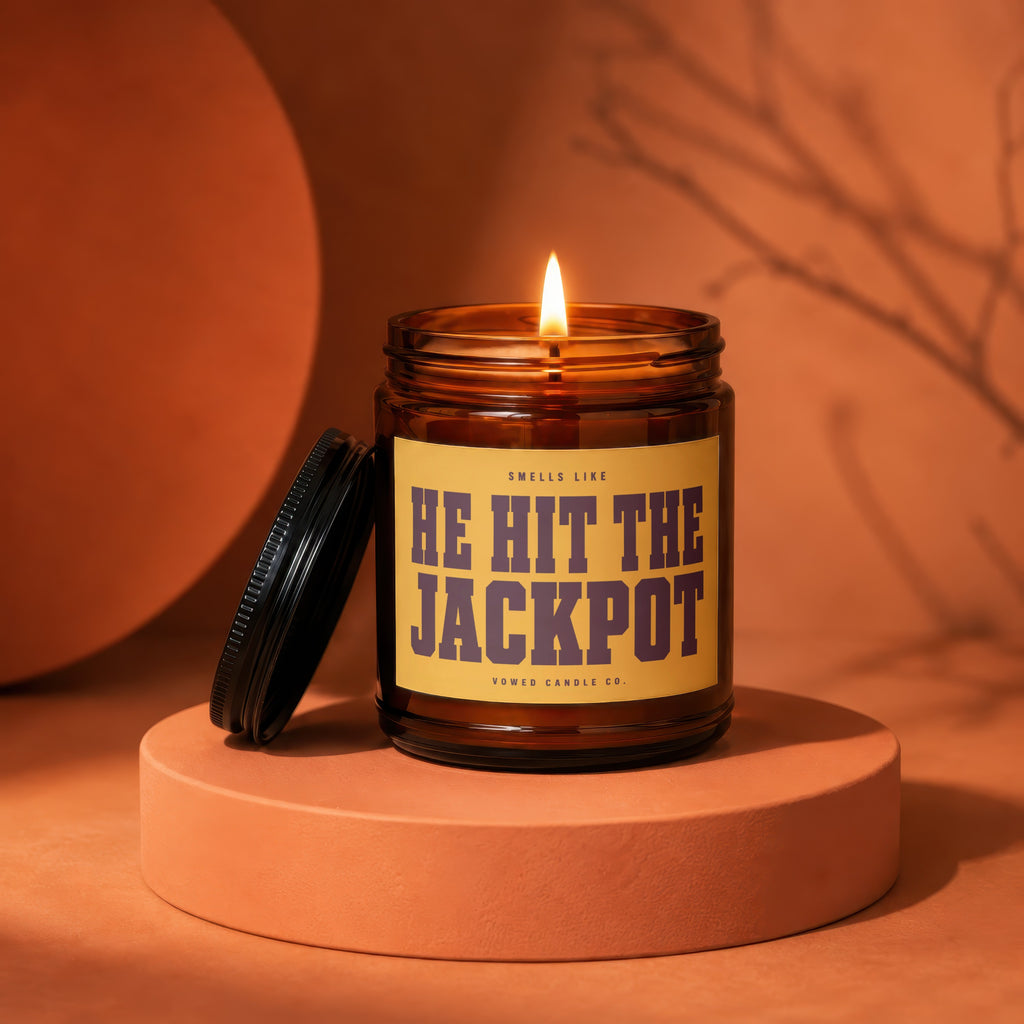 Amber candle with lit wick, label reads "HE HIT THE JACKPOT".