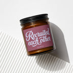 Amber jar candle with maroon label that reads "Recruited each other, VOWED."