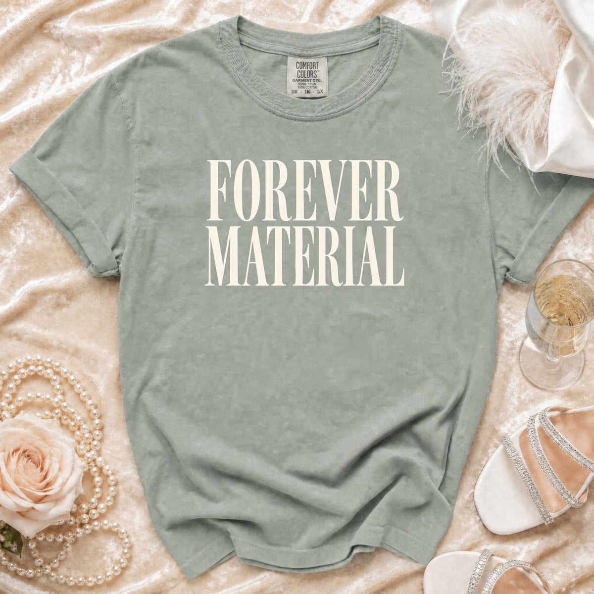 Sage green t-shirt with "FOREVER MATERIAL" text, styled with pearls and champagne.
