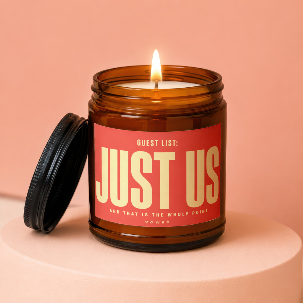 Amber jar candle with "JUST US" label and lit flame on a peach-colored pedestal.