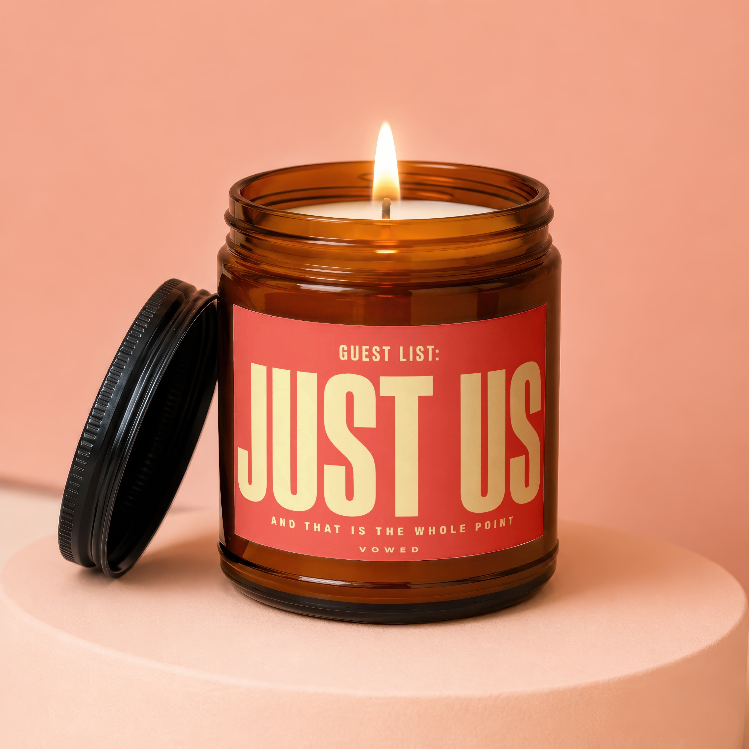 Amber jar candle with "JUST US" label and lit flame on a peach-colored pedestal.