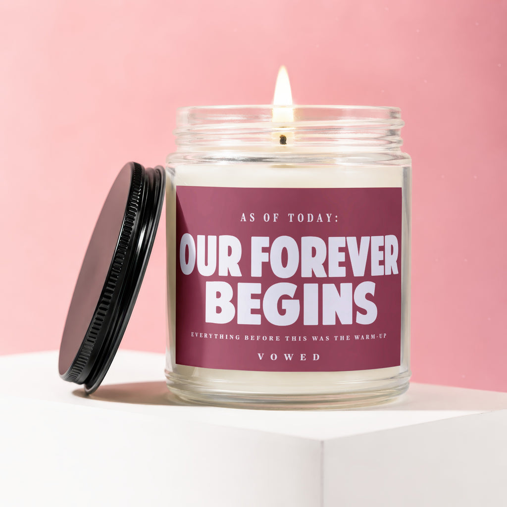 Candle with text "Our Forever Begins" on a pink background, black lid beside it.