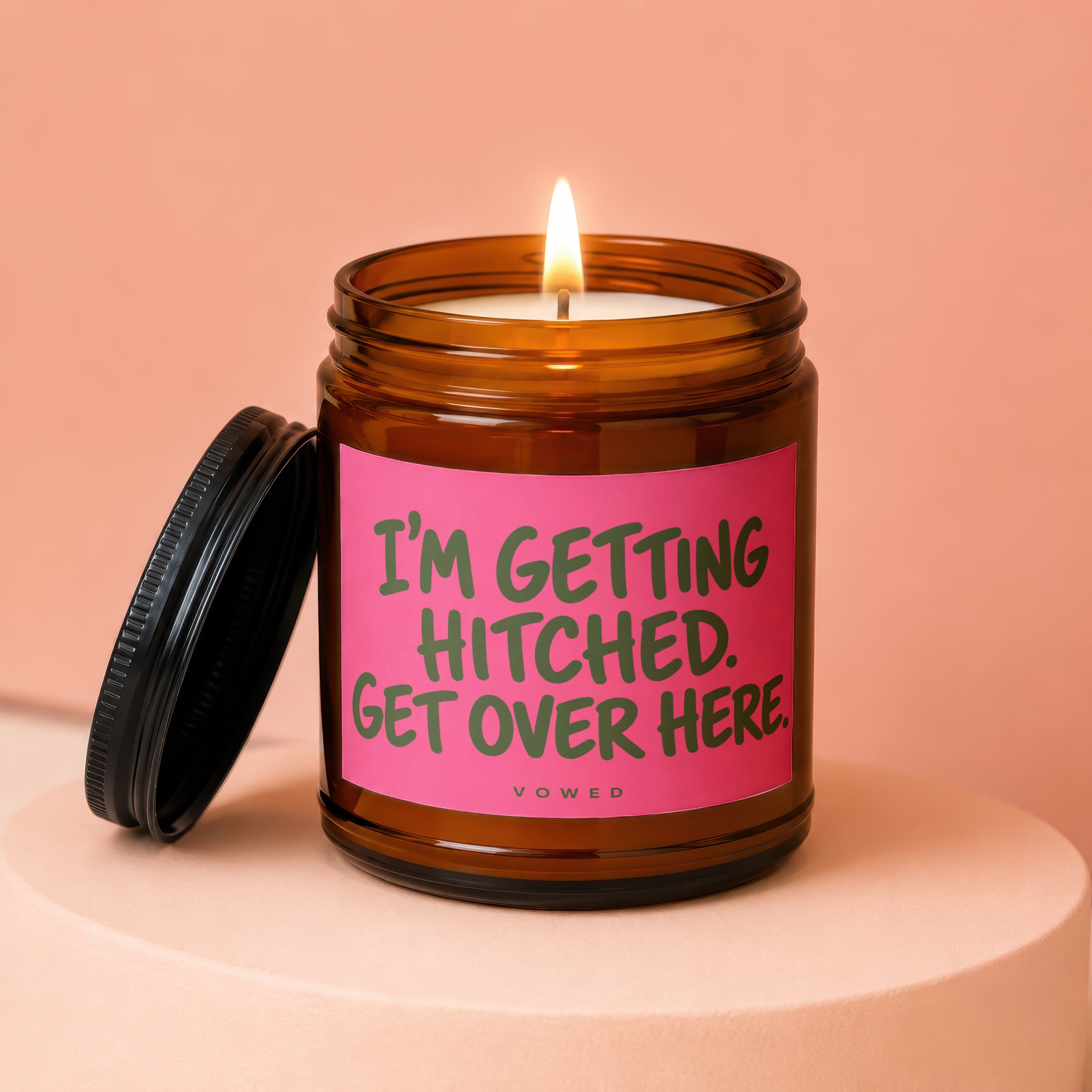Amber candle in jar with lit wick and black lid, featuring pink label with green text.