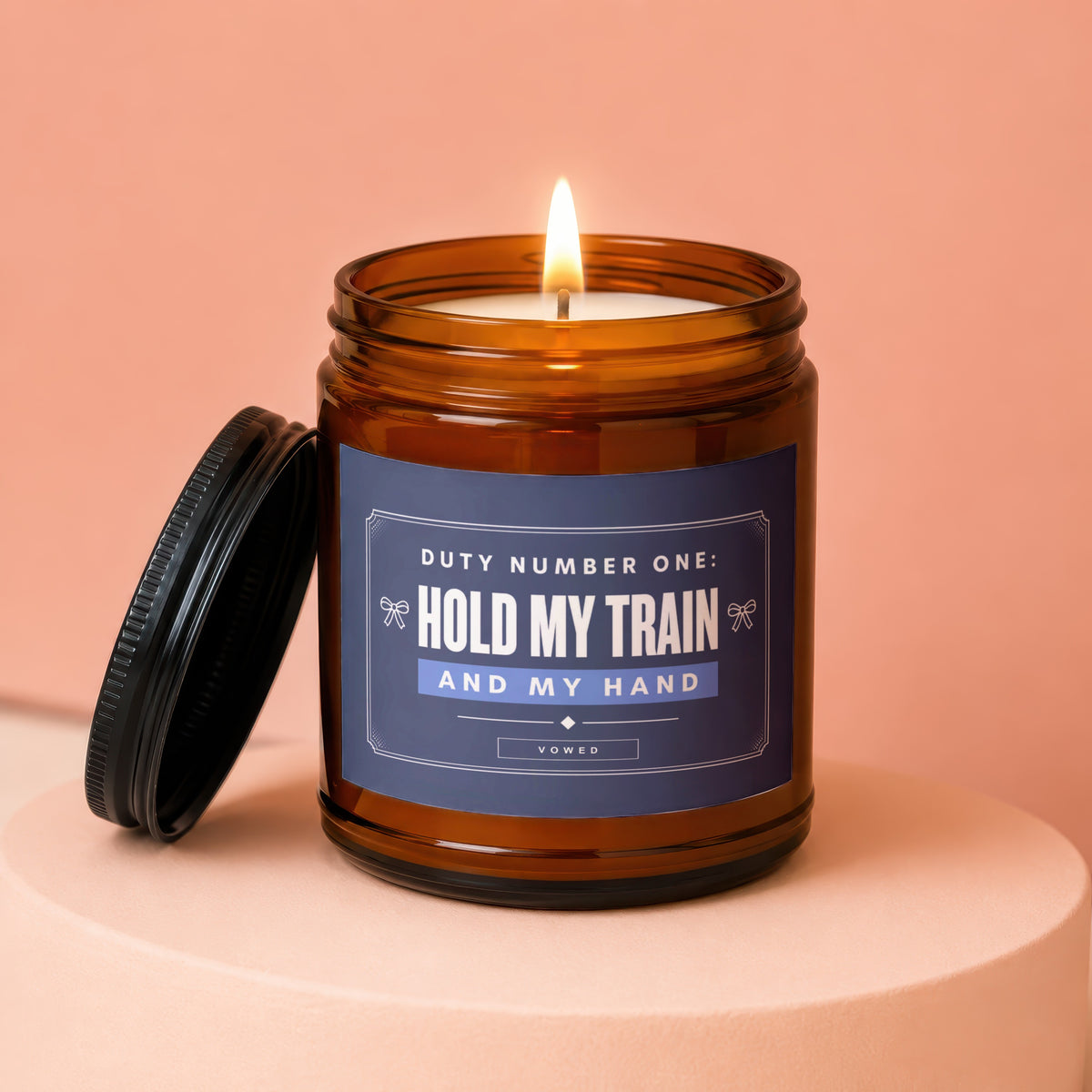 Amber candle with lit wick, lid off, text "Hold My Train And My Hand".