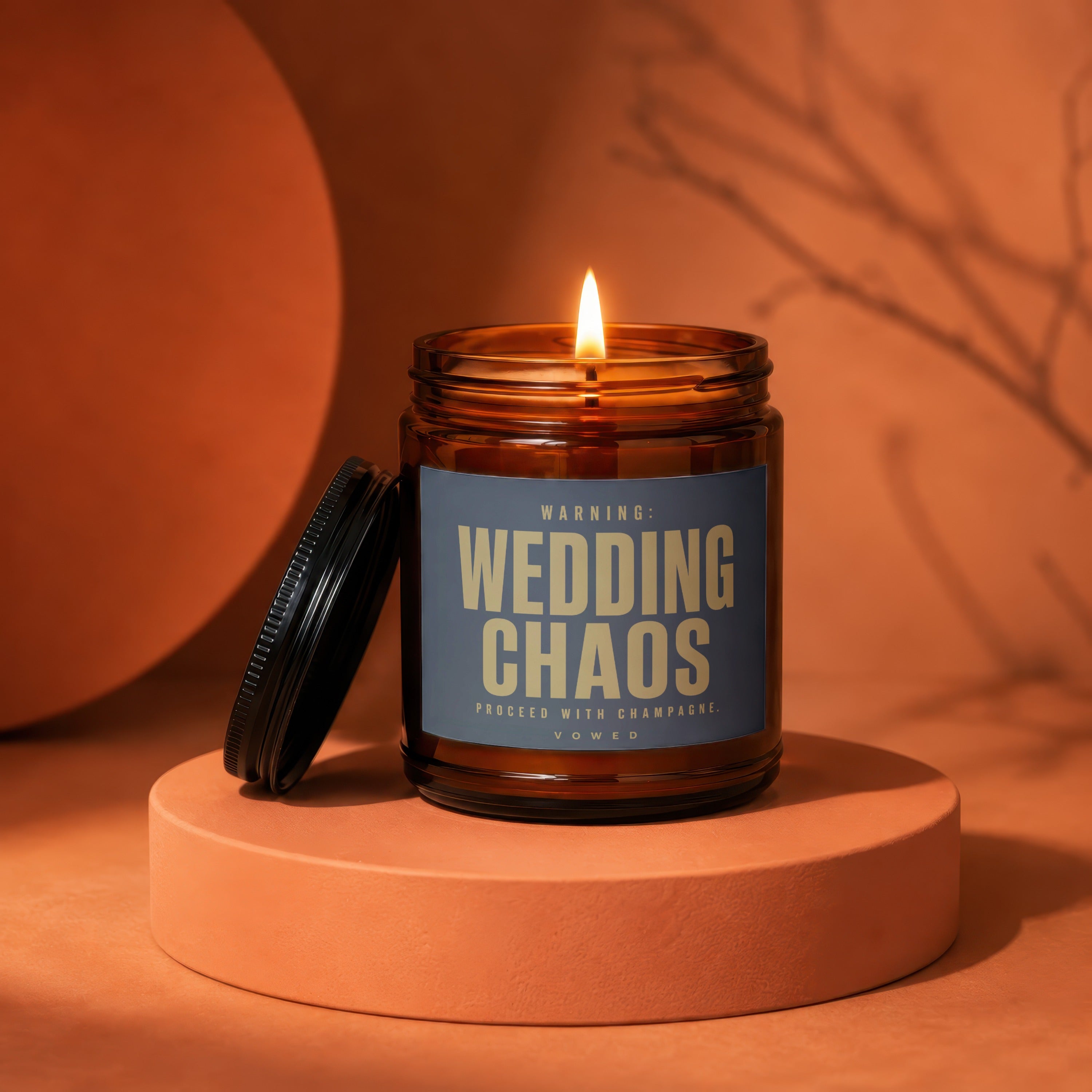 Amber jar candle with lit flame, features "WEDDING CHAOS. PROCEED WITH CHAMPAGNE. VOWED" text.