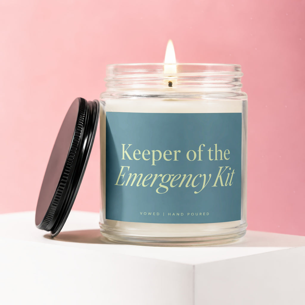 Glass jar candle with "Keeper of the Emergency Kit" text and lit wick.