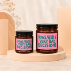 Two amber glass candles with pink labels featuring teal text: "VOWS, VEILS & VERY BAD DECISIONS".
