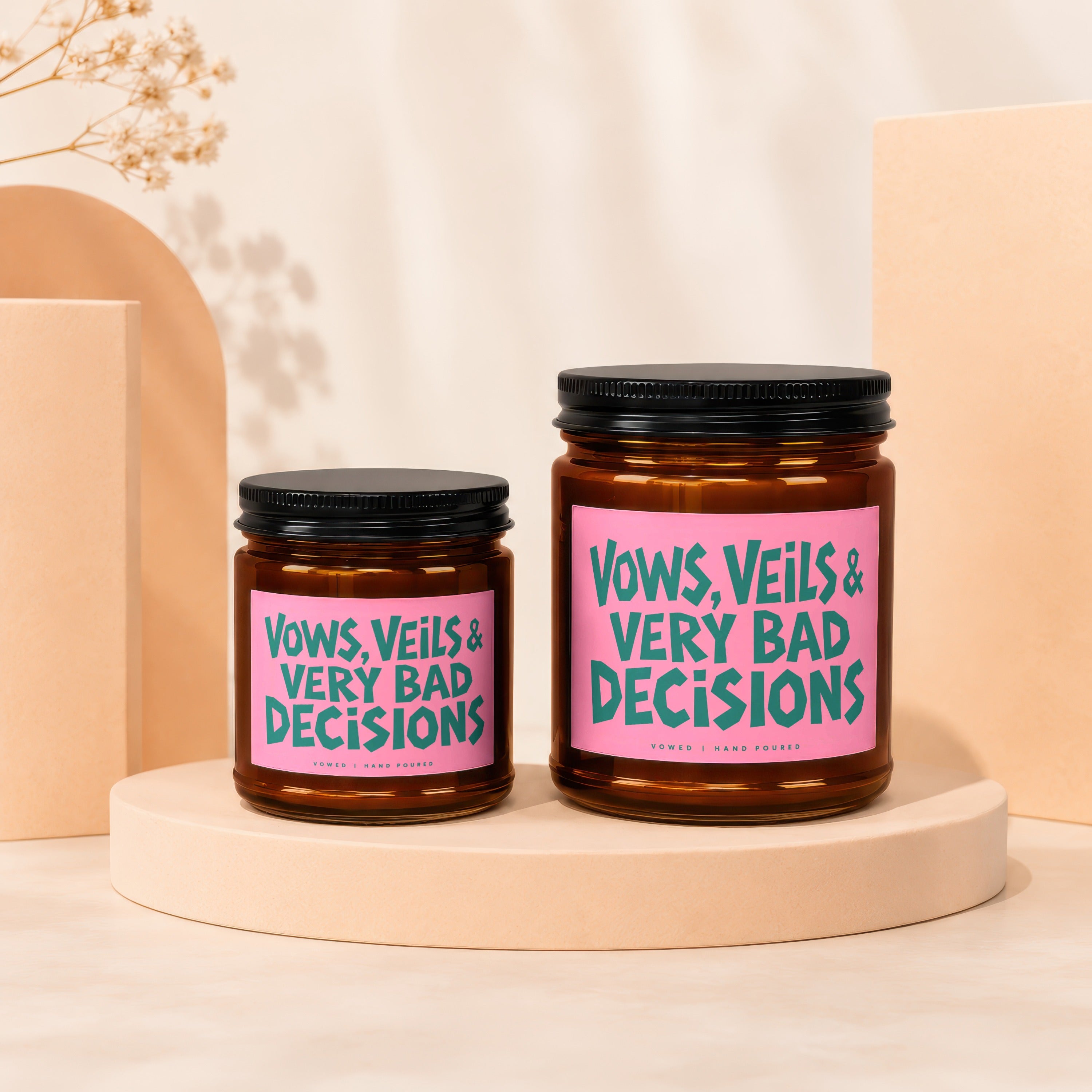 Two amber glass candles with pink labels featuring teal text: "VOWS, VEILS & VERY BAD DECISIONS".