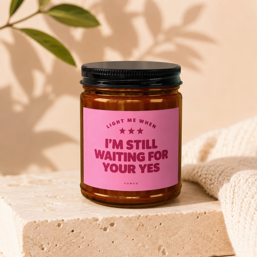 Amber candle with pink label reading "Light me when I'm still waiting for your yes".