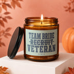 Amber candle with lit wick and label reading "TEAM BRIDE RECRUIT VETERAN VOWED".