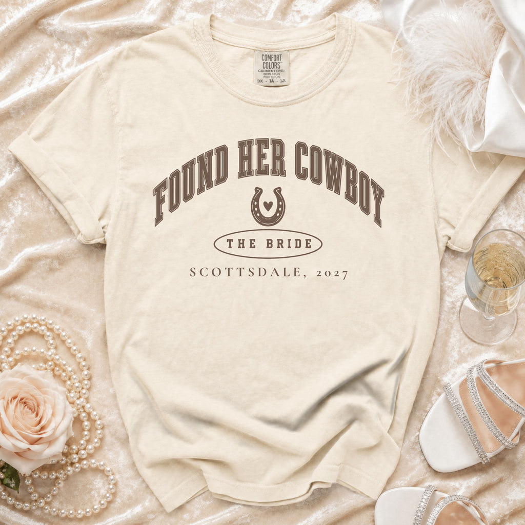 Cream t-shirt with brown text reading FOUND HER COWBOY THE BRIDE SCOTTSDALE, 2027, on textured fabric.