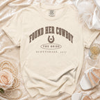 Cream t-shirt with brown text reading FOUND HER COWBOY THE BRIDE SCOTTSDALE, 2027, on textured fabric.