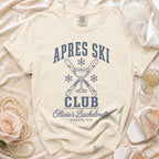 Cream t-shirt featuring blue Apres Ski Club text, crossed skis, snowflakes, and champagne glass graphic.