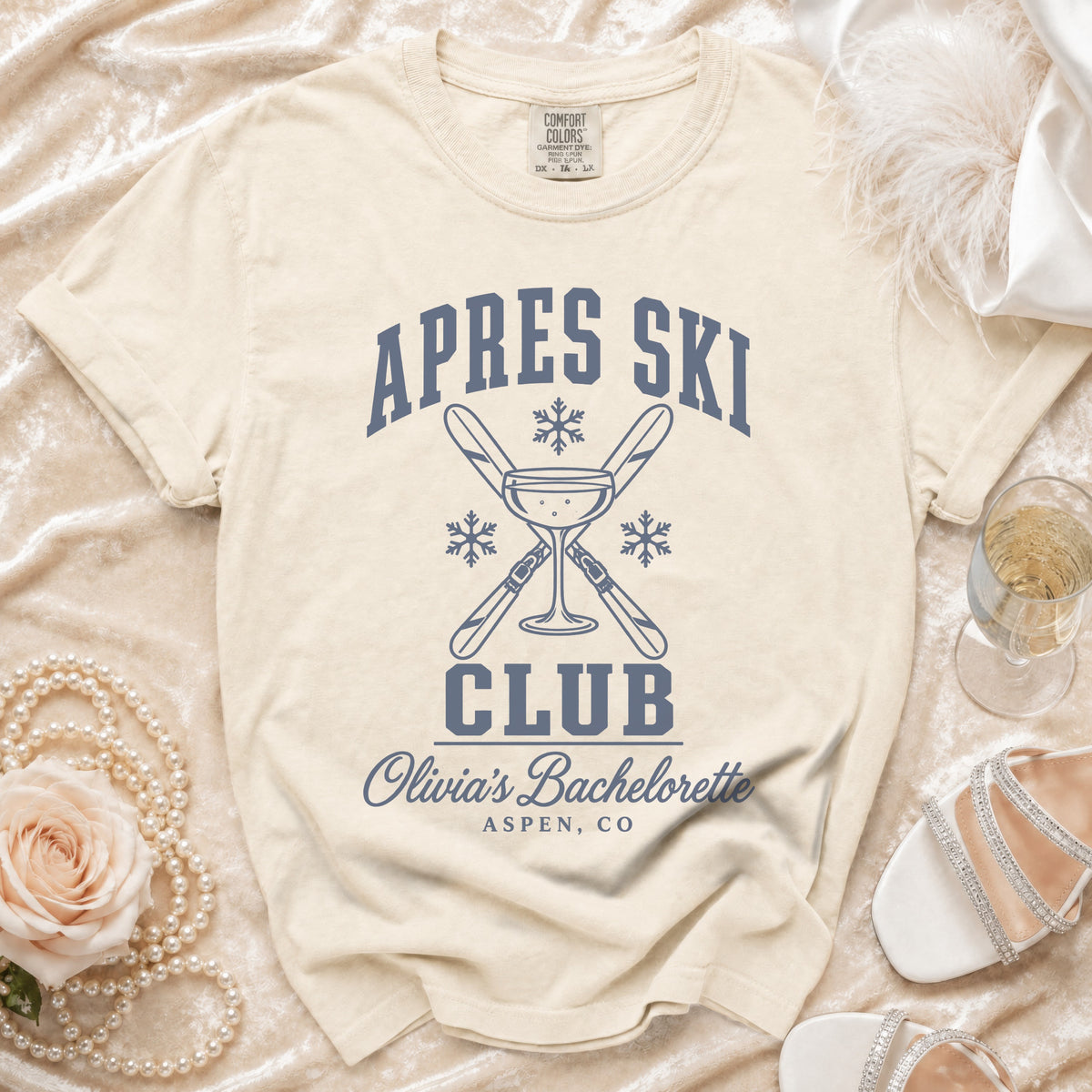 Cream t-shirt featuring blue Apres Ski Club text, crossed skis, snowflakes, and champagne glass graphic.