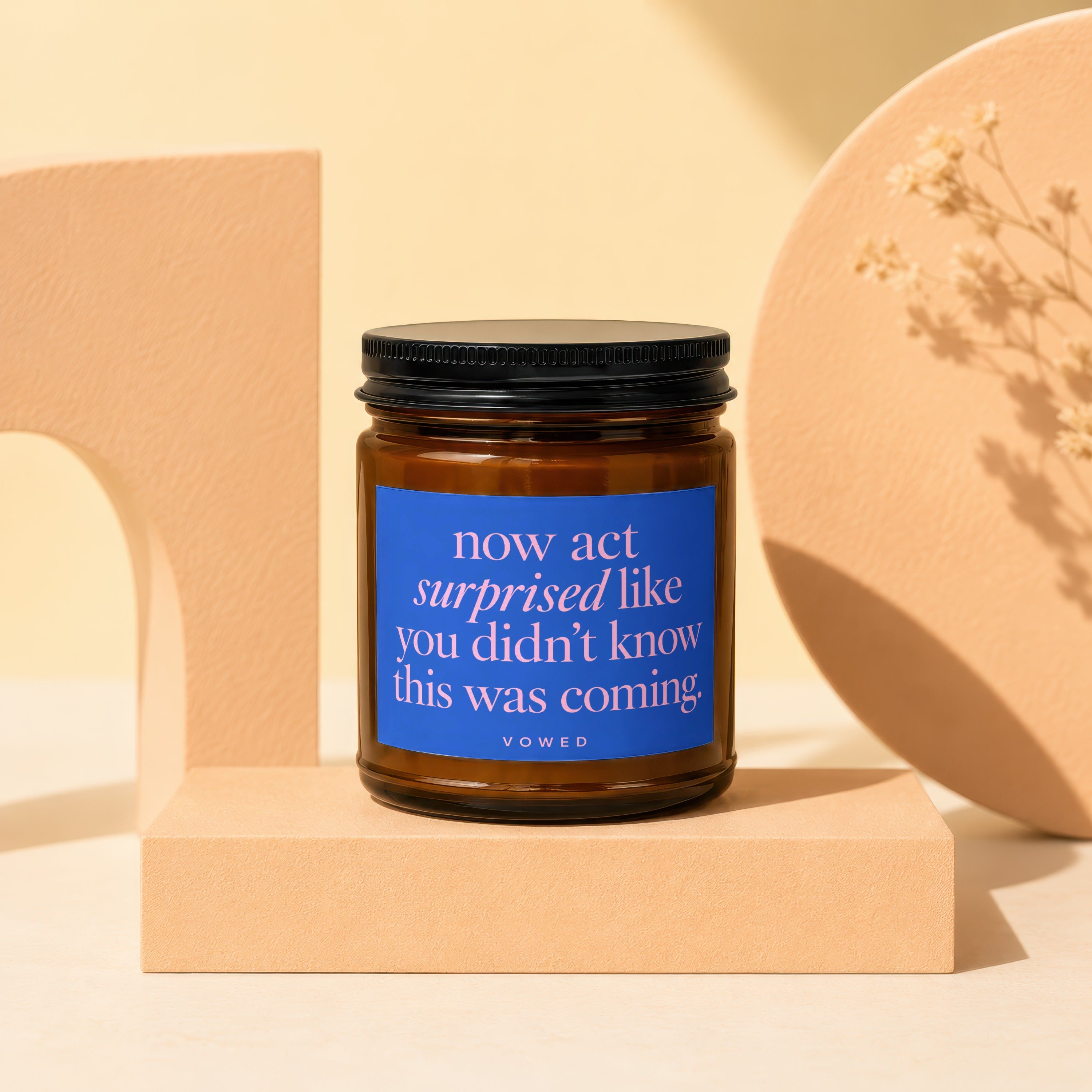 Brown glass candle with a blue label reading "now act surprised like you didn't know this was coming."
