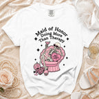 White t-shirt with graphic of skeleton in chair, holding drink, with text "Maid of Honor Doing More Than Therapy".