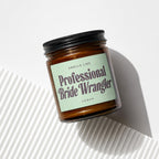 Amber glass candle with black lid, light green label that reads "Smells Like Professional Bride Wrangler Vowed".