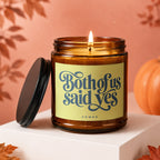 Amber glass candle with lit wick and "Both of us said yes VOWED" text on label.