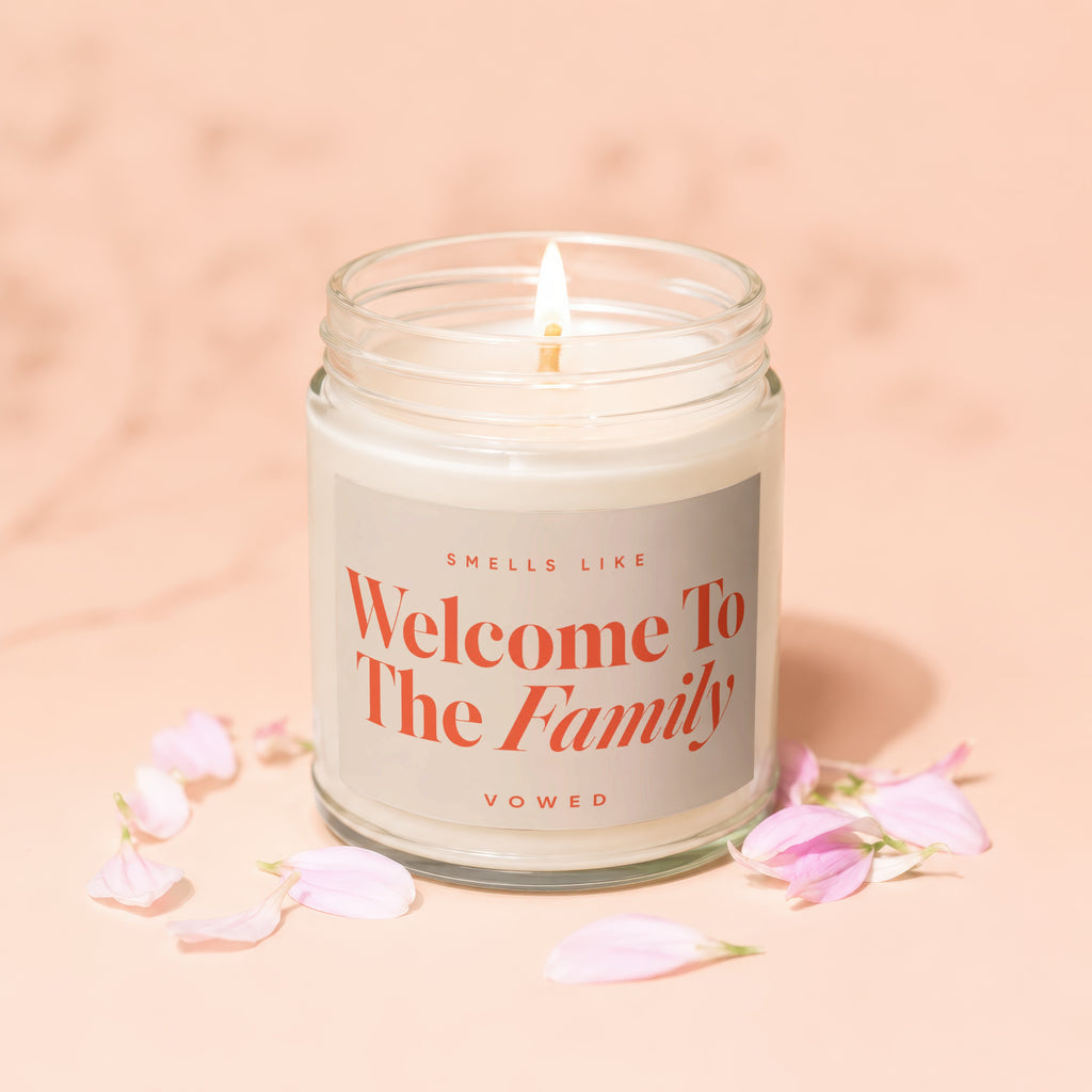 Glass jar candle with lit wick. Label reads 'Smells Like Welcome To The Family'.