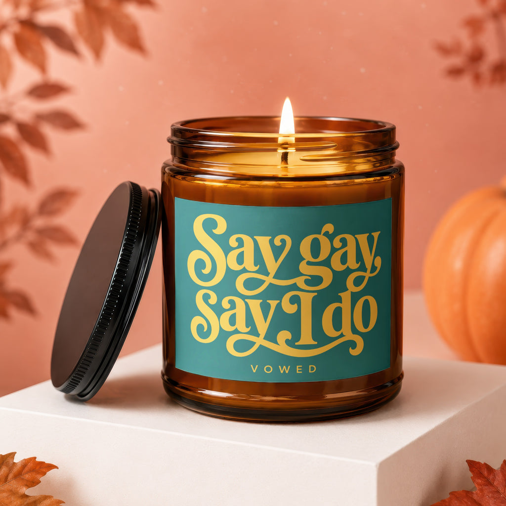 Amber candle with burning wick, lid off, featuring teal label with gold text "Say gay Say I do".