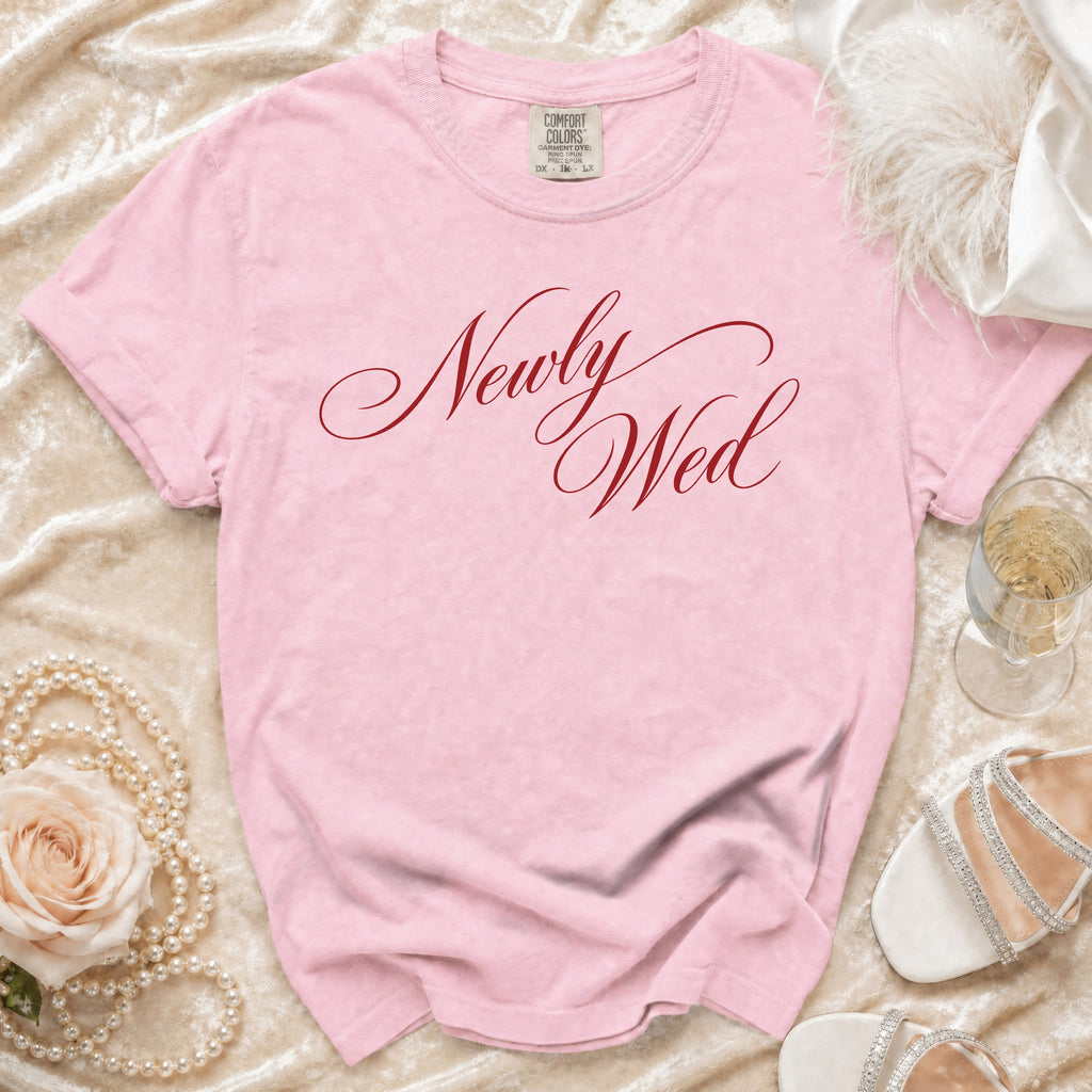 Pink t-shirt with red script text "Newly Wed," pearl necklace, and white heels.