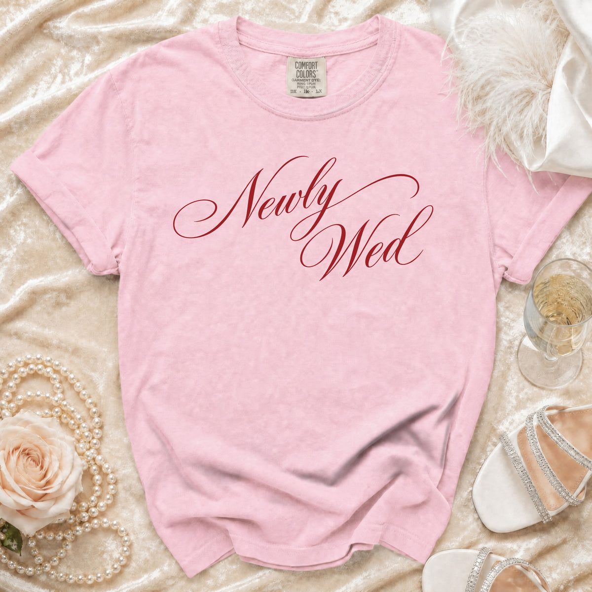 Pink t-shirt with red script text "Newly Wed," pearl necklace, and white heels.