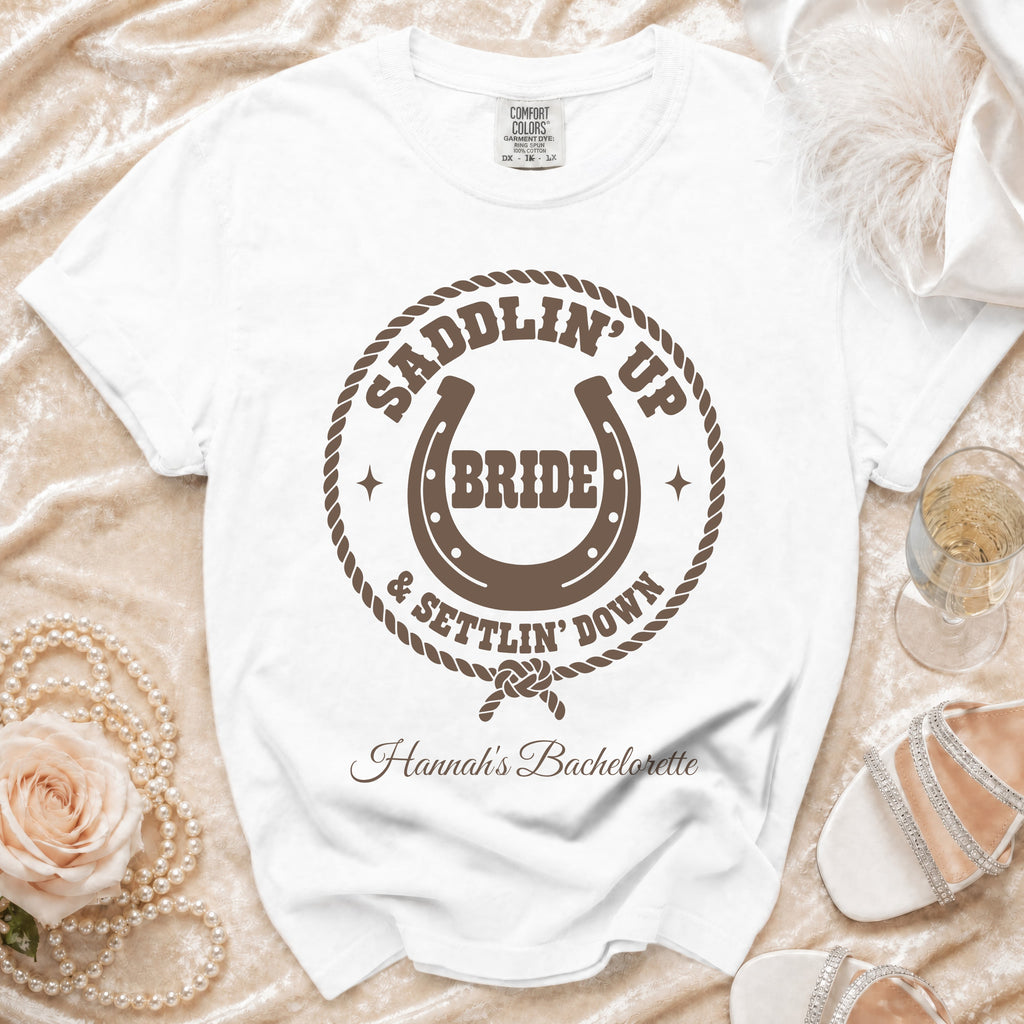 A white t-shirt featuring brown western-themed text, a horseshoe, and rope border, laid on a satin background with a rose and pearl necklace.