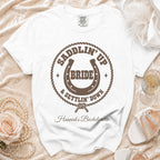 A white t-shirt featuring brown western-themed text, a horseshoe, and rope border, laid on a satin background with a rose and pearl necklace.