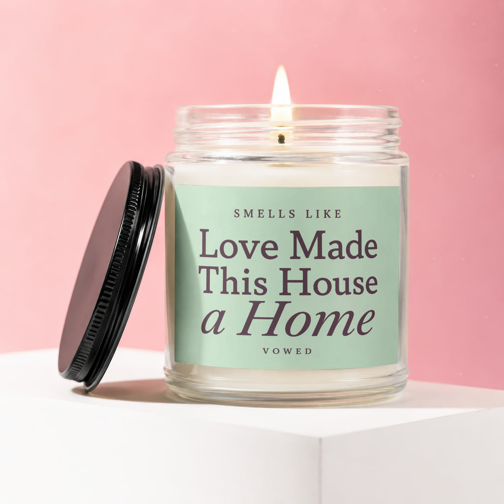 Lit candle in a glass jar with lid off, reading "Smells Like Love Made This House a Home".